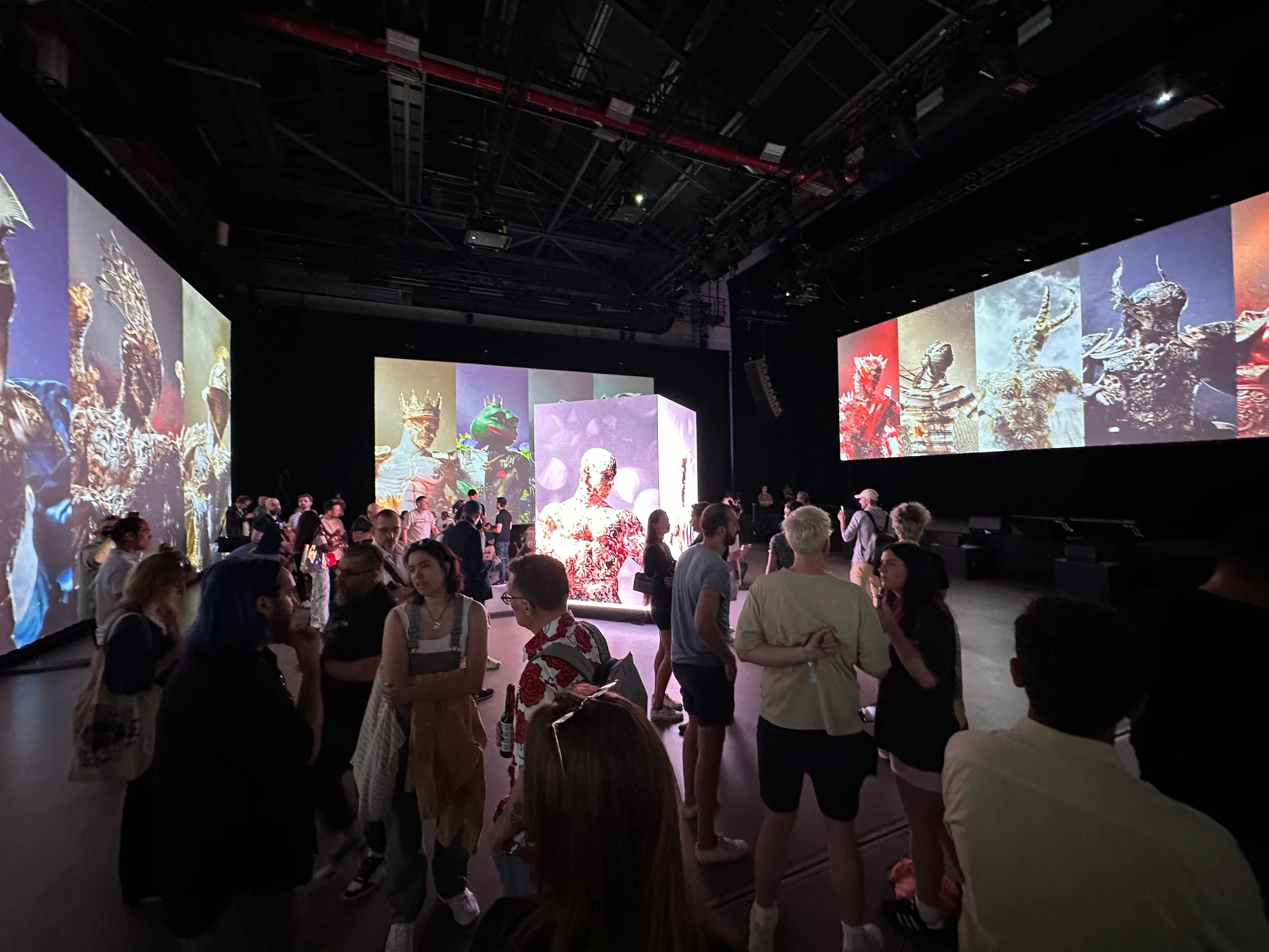 A dark hall with Nexus Anima portraits projected across the walls and a lit screen cube, visitors in between