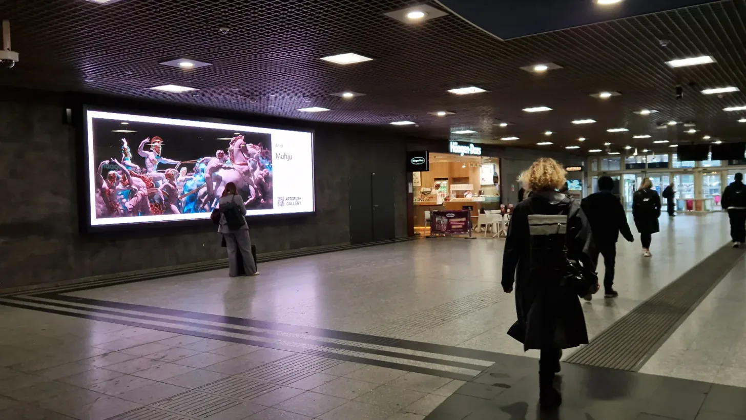 People pass a long digital display showing a Muhju artwork in a Milan metro station passage.