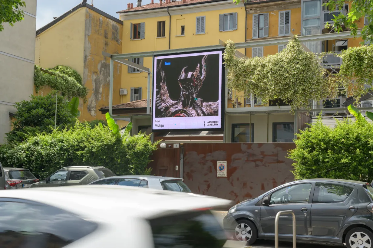 An outdoor digital screen displays a Muhju artwork among trees and yellow buildings on a Milan street.