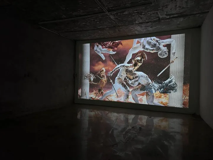 A projected battle scene with winged figures reflects on the wet floor of a dark concrete room.