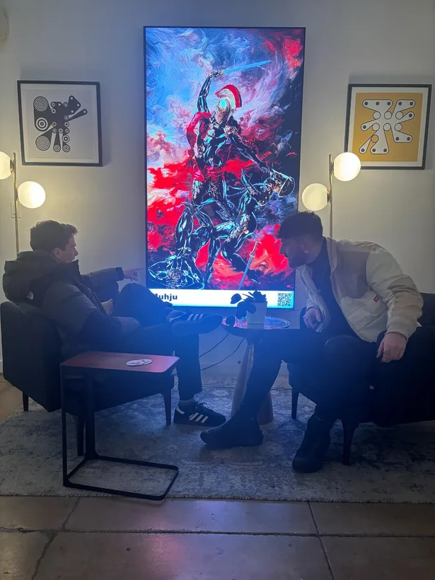Two visitors sit in a lounge before a tall screen displaying a red and blue armoured warrior.