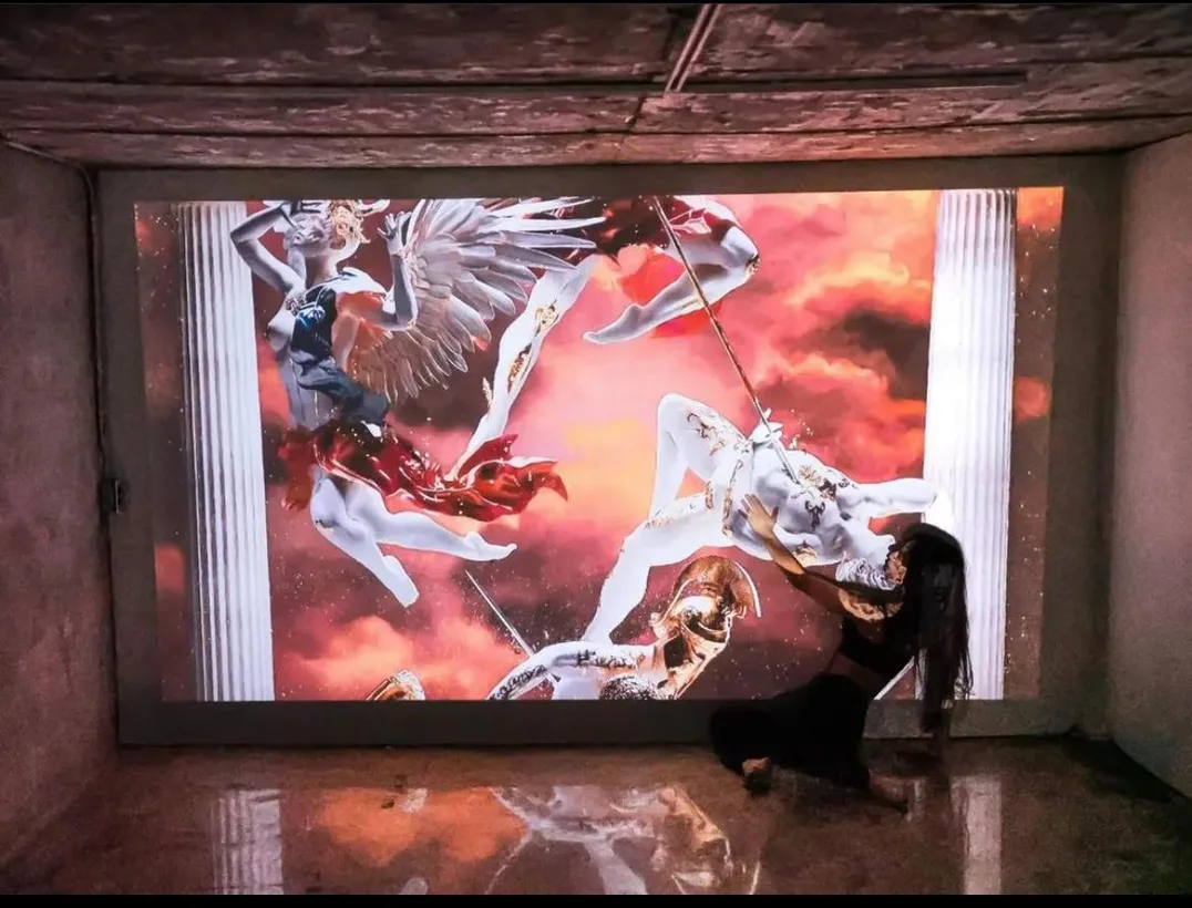 A dancer kneels before a projected battle scene with winged figures in a concrete room.
