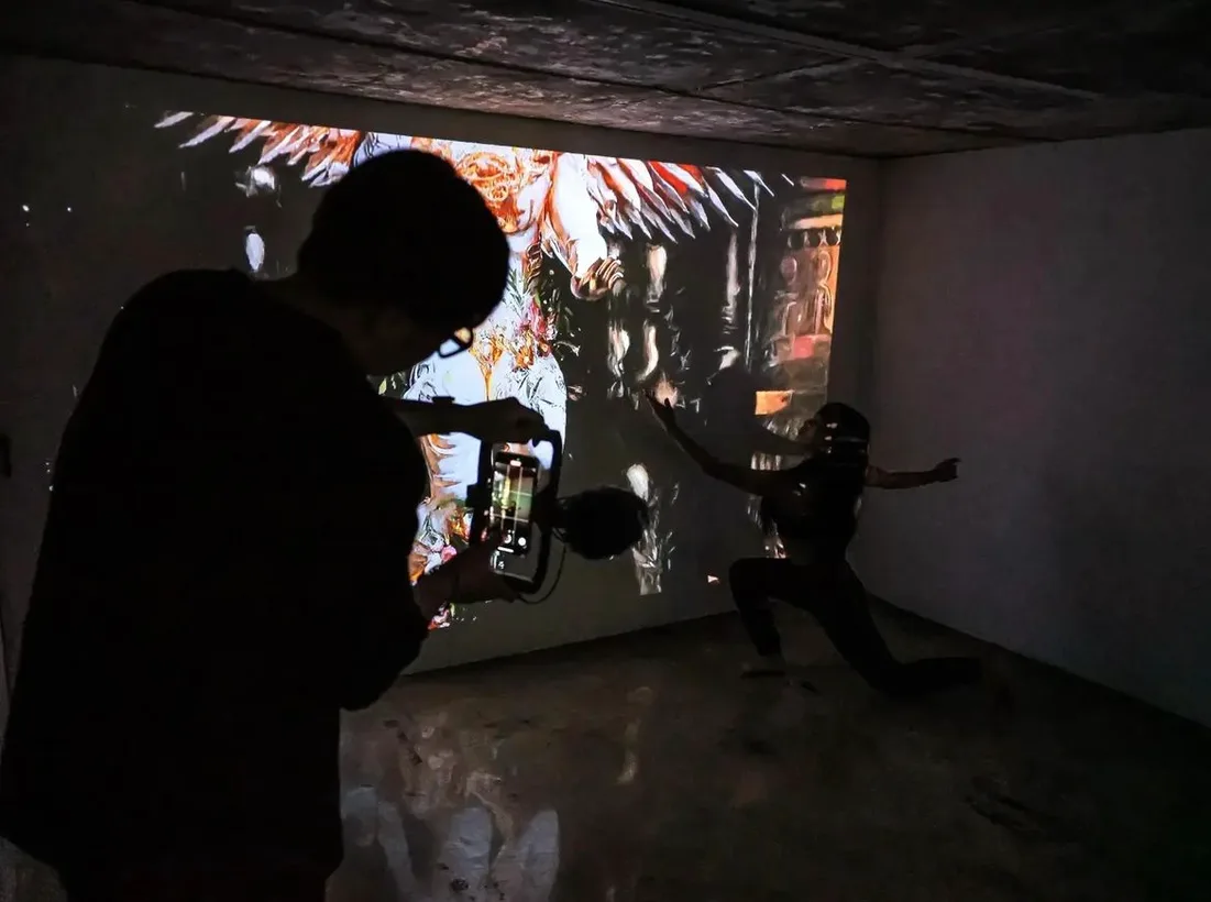 A man films a performer dancing before a projected Muhju artwork in a dark room.