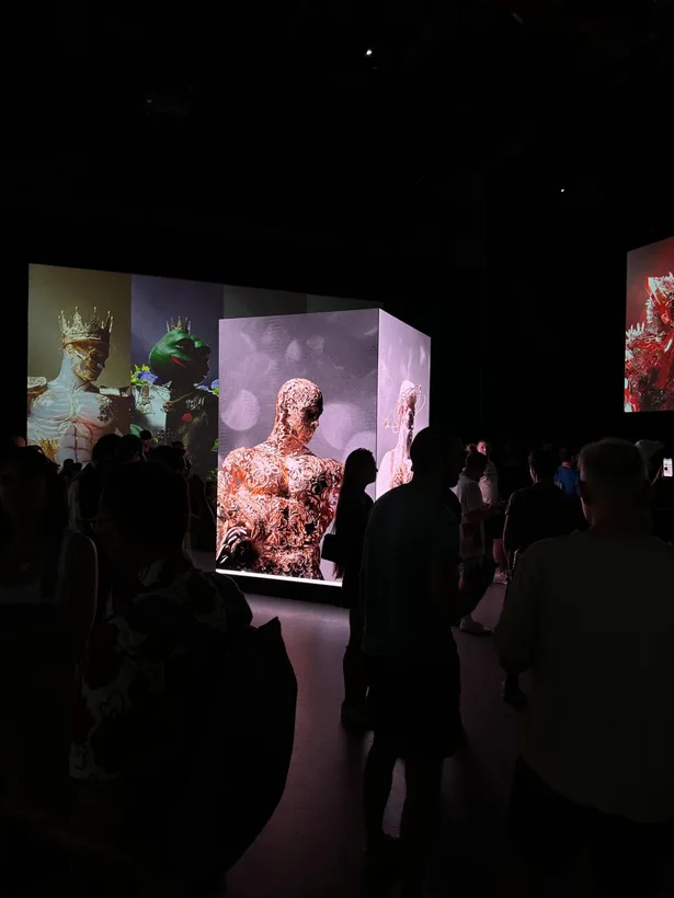 Visitors stand in silhouette before a screen displaying a sculpted figure with red veins.