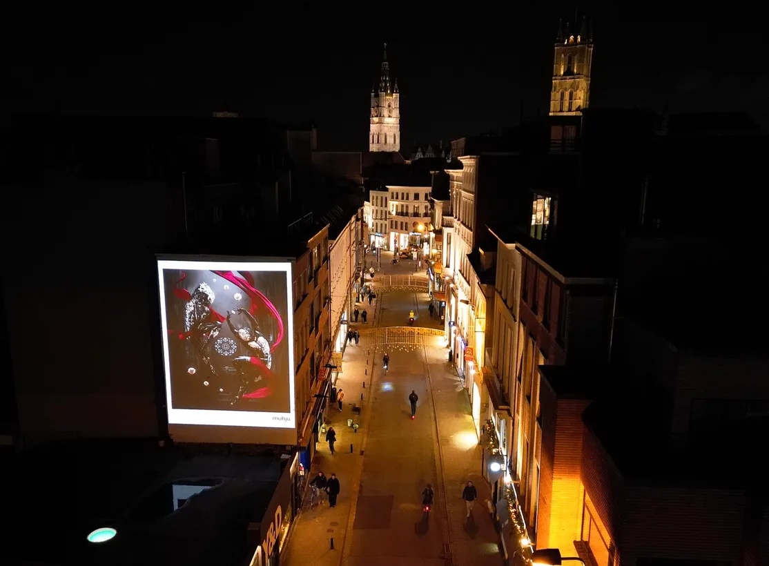 An illuminated billboard displays a Muhju artwork above a narrow European street at night.
