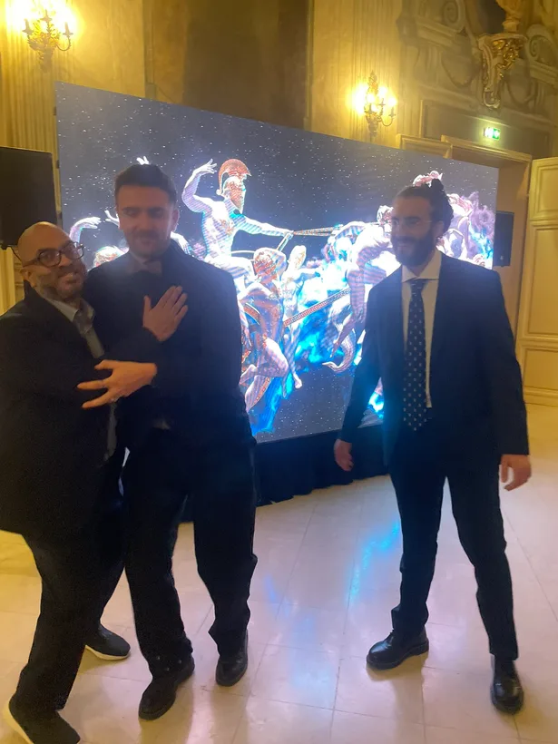 Three visitors in suits stand before a screen displaying a colourful baroque scene in an ornate hall.