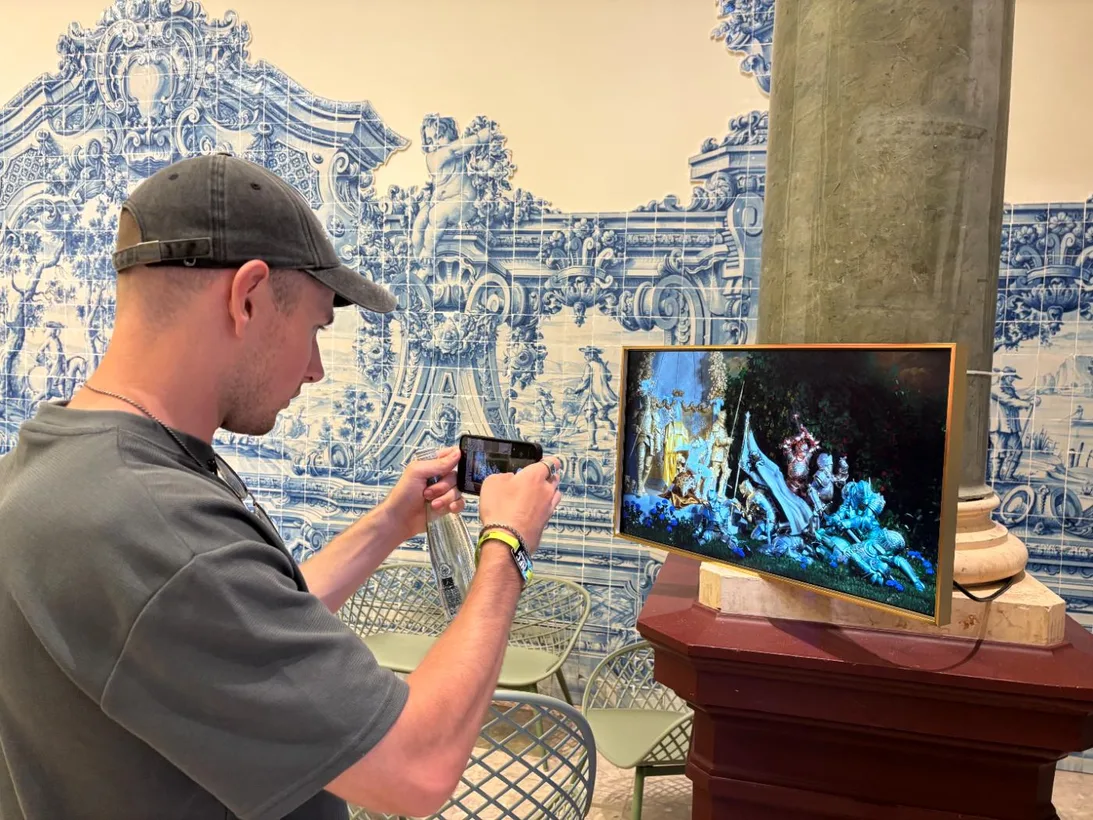 A man photographs a framed Muhju artwork on a red pedestal before blue and white Lisbon tiles.