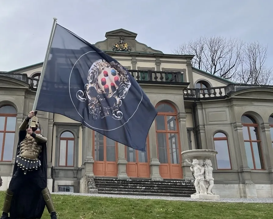 An armoured performer raises a black flag bearing Muhju's skull emblem before a neoclassical villa.