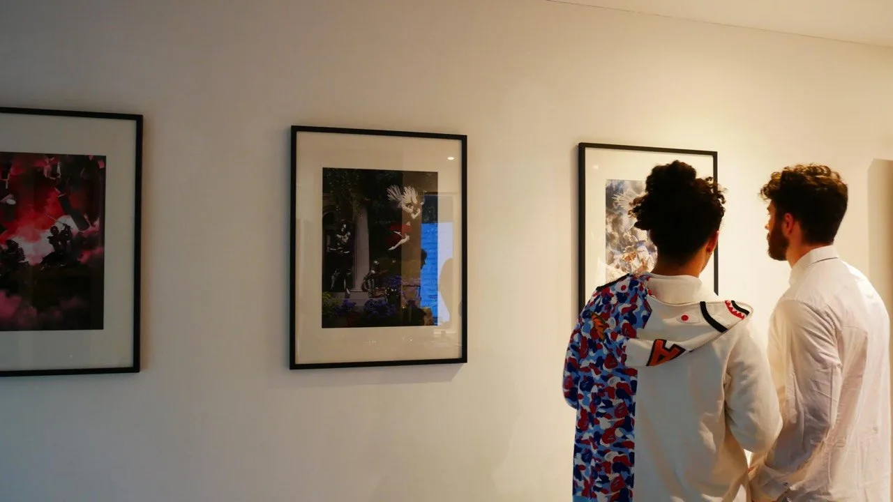 Two visitors look at framed Muhju artworks on a light gallery wall.