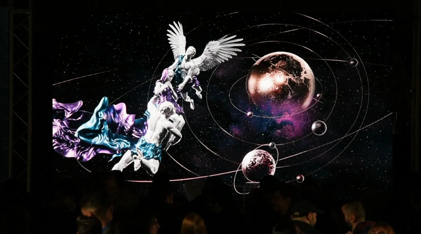 A huge screen above an audience displays a Muhju artwork featuring a winged figure among orbiting planets.