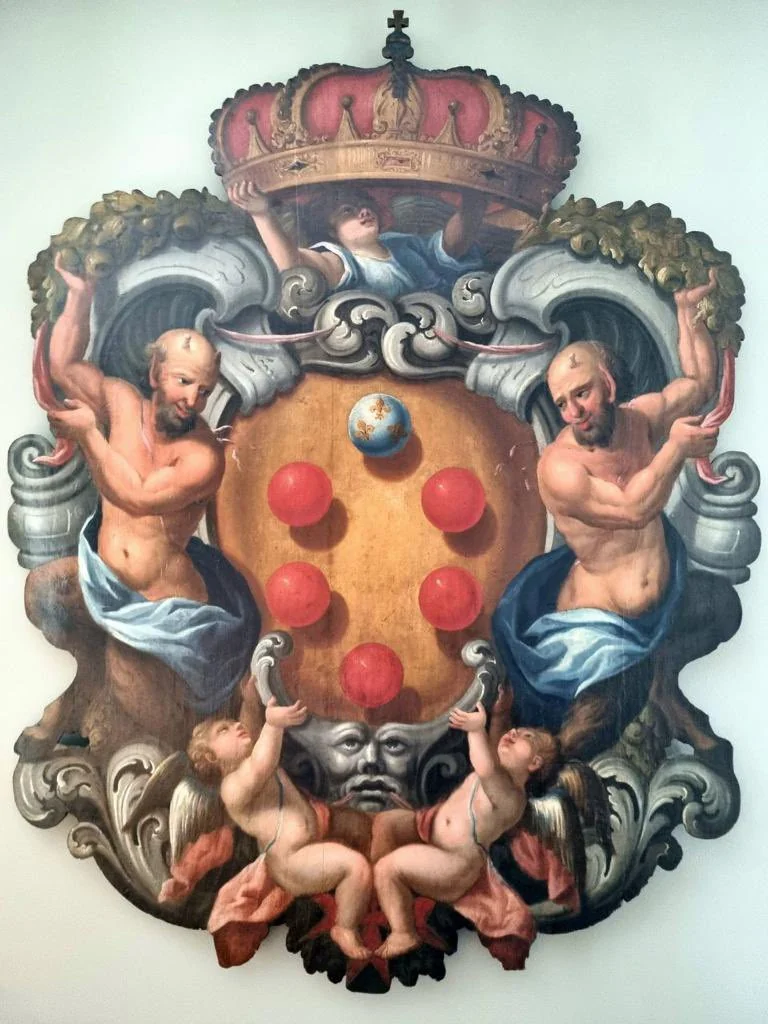 Painted Medici arms: a golden shield with red balls and a blue orb beneath a crown, flanked by two muscular nude supporters with putti below.